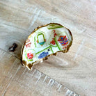 A handmade ring shaped like a broken shell, featuring a white enamel face decorated with colorful Chinese-style tiles, characters, and motifs, and finished with a gold-tone rim, resting on a light wood surface.