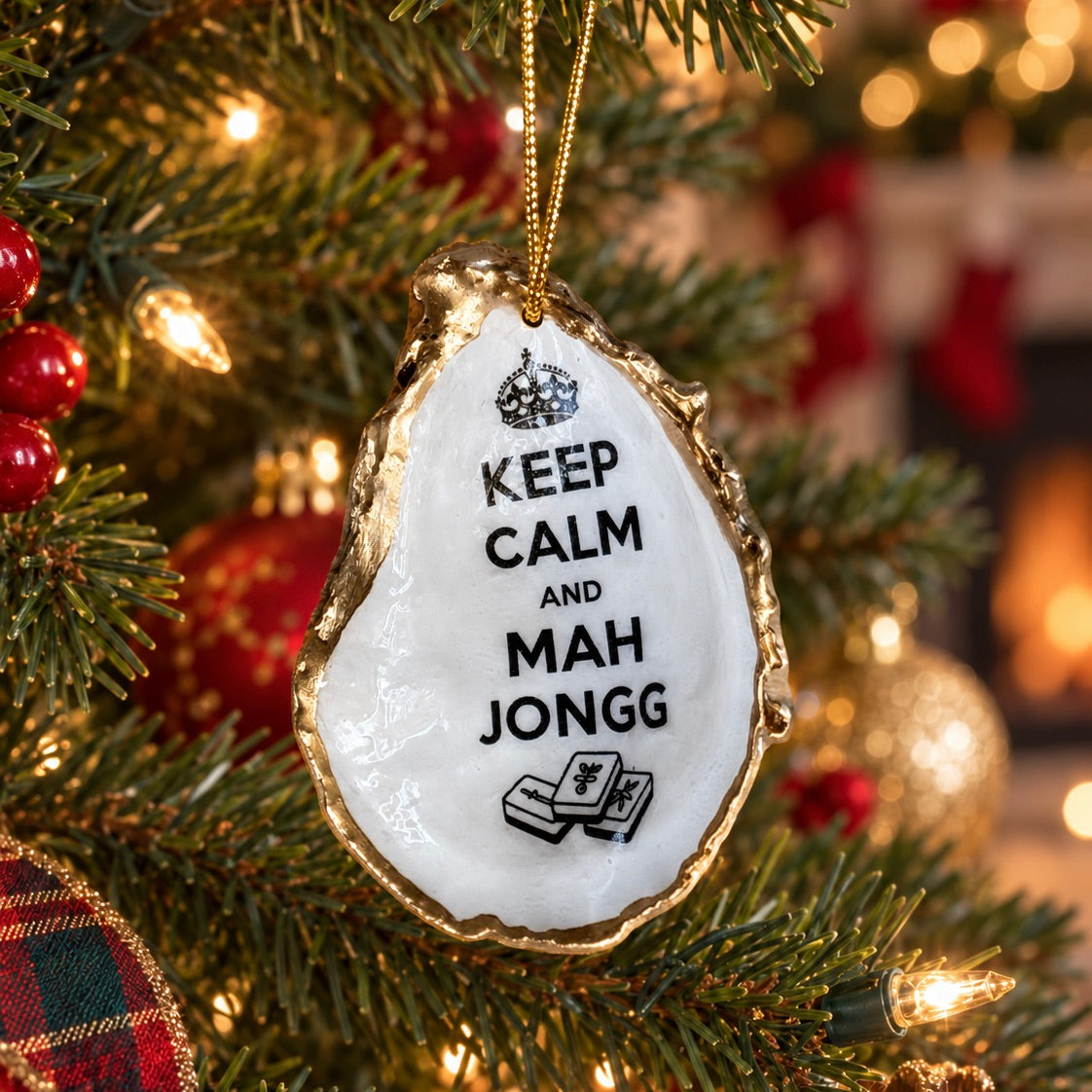 A ceramic Christmas ornament shaped like an oyster shell hangs on a decorated Christmas tree. The ornament is white with a gold-toned irregular edge, black text reading 'KEEP CALM AND MAH JONGG', a small crown graphic at the top, and a small black illustration at the bottom.