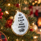 A ceramic Christmas ornament shaped like an oyster shell hangs on a decorated Christmas tree. The ornament is white with a gold-toned irregular edge, black text reading 'KEEP CALM AND MAH JONGG', a small crown graphic at the top, and a small black illustration at the bottom.