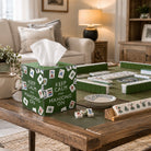 A green Mahjong-themed tissue box cover sits on a wooden table in a cozy living room, with a mahjong game set arranged on the tabletop nearby.