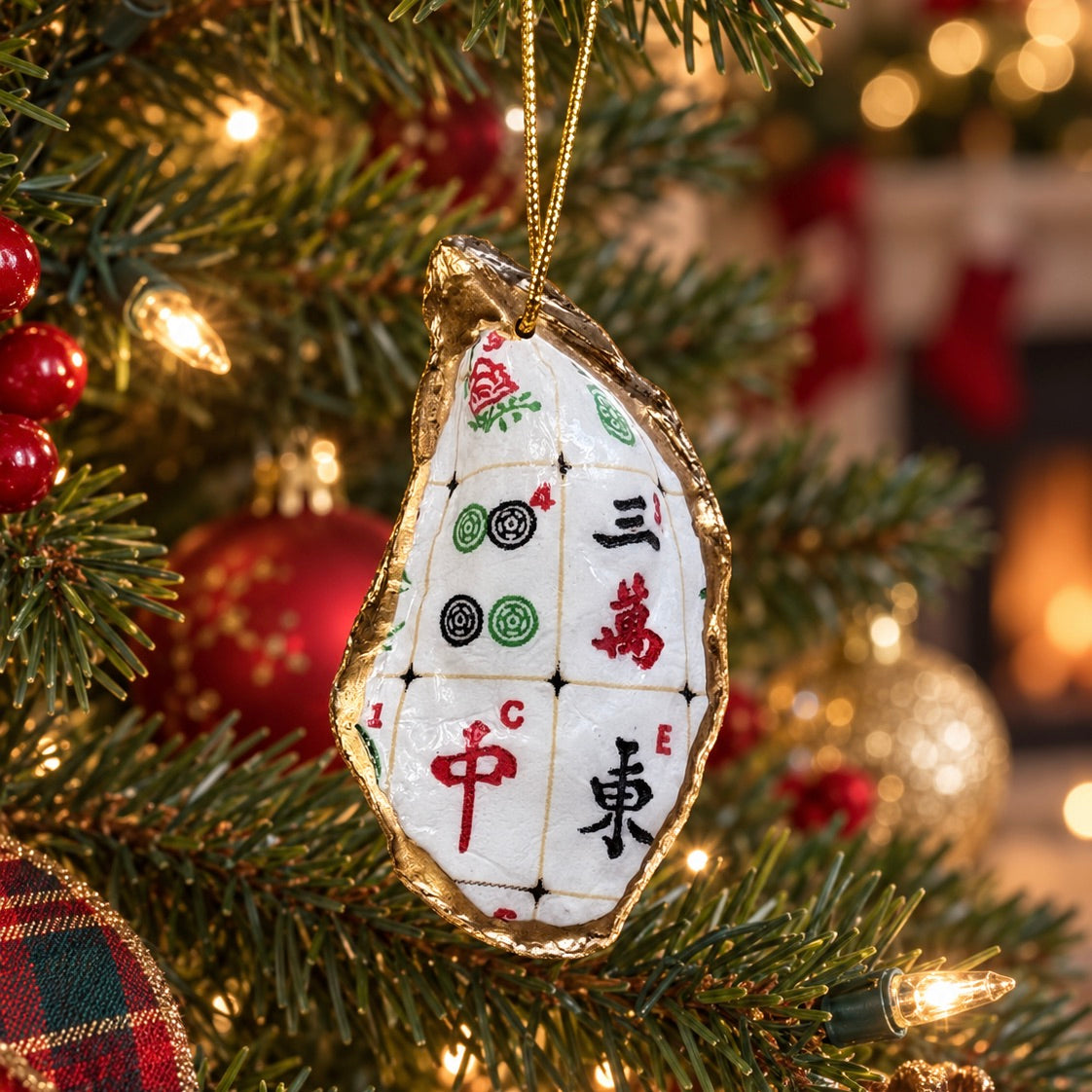 A decorative Christmas ornament shaped like an oyster shell, featuring a white mahjong tile design with red, green, and black symbols and gold foil edges, hanging from a lit Christmas tree.