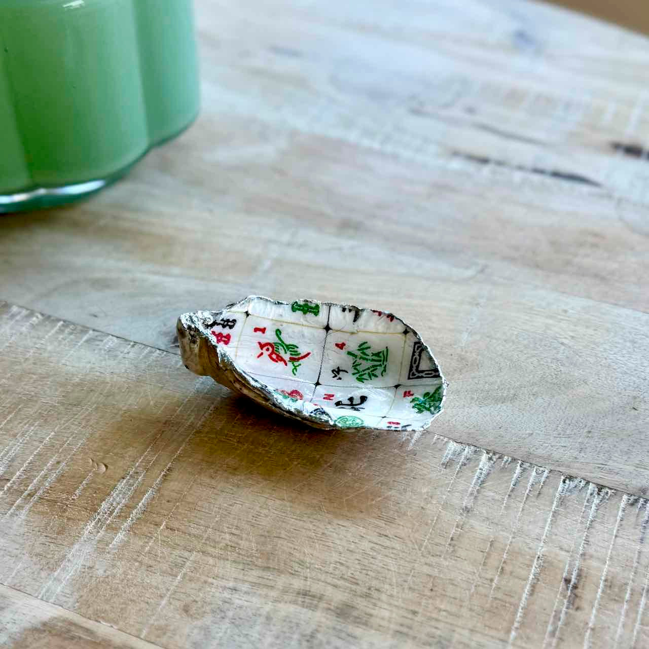 A small decorative shell-shaped dish with a white background and a printed pattern of green pine trees and red ornaments, resting on a light wood tabletop.