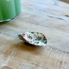A small decorative shell-shaped dish with a white background and a printed pattern of green pine trees and red ornaments, resting on a light wood tabletop.