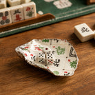 A small decorative dish shaped like an open shell, printed with a mahjong board design and holding two white dice, set on a wooden tabletop with mahjong tiles nearby.