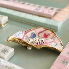 A decorative shell-shaped dish with gold-edged trim and a pink floral pattern, holding three white dice, set on a mint green tabletop with mahjong tiles in the background.