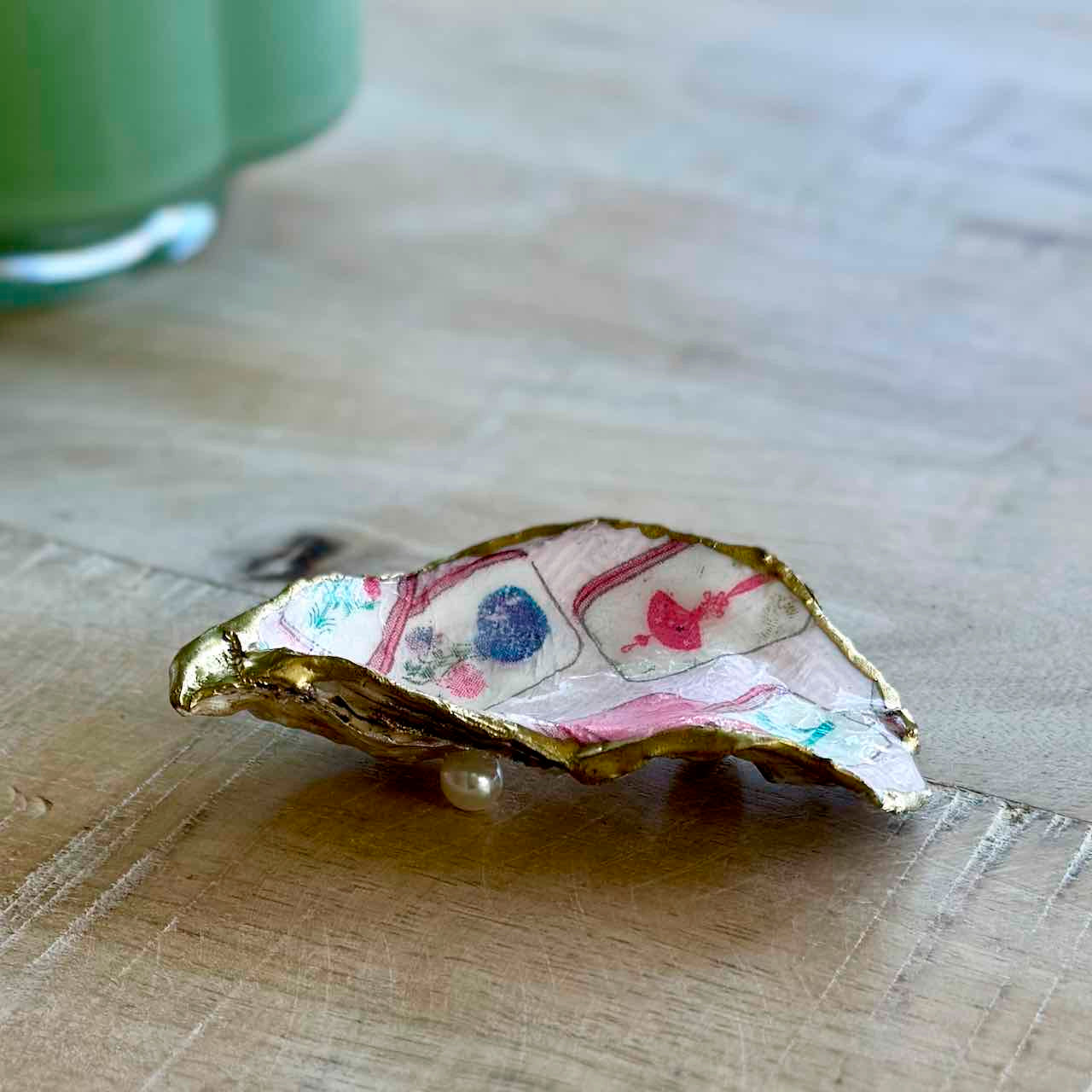 A small decorative shell-shaped object with a gold-edged rim and a white interior printed with pink playing card suits and blue floral details, resting on a light wood surface.