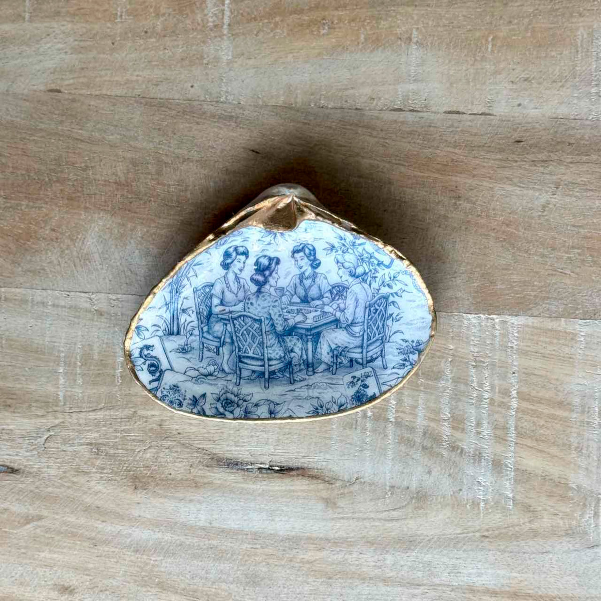 A small shell-shaped decorative object with a blue and white illustrated scene on one side and a gold-toned rim, photographed on a light wood surface.
