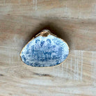 A small shell-shaped decorative object with a blue and white illustrated scene on one side and a gold-toned rim, photographed on a light wood surface.