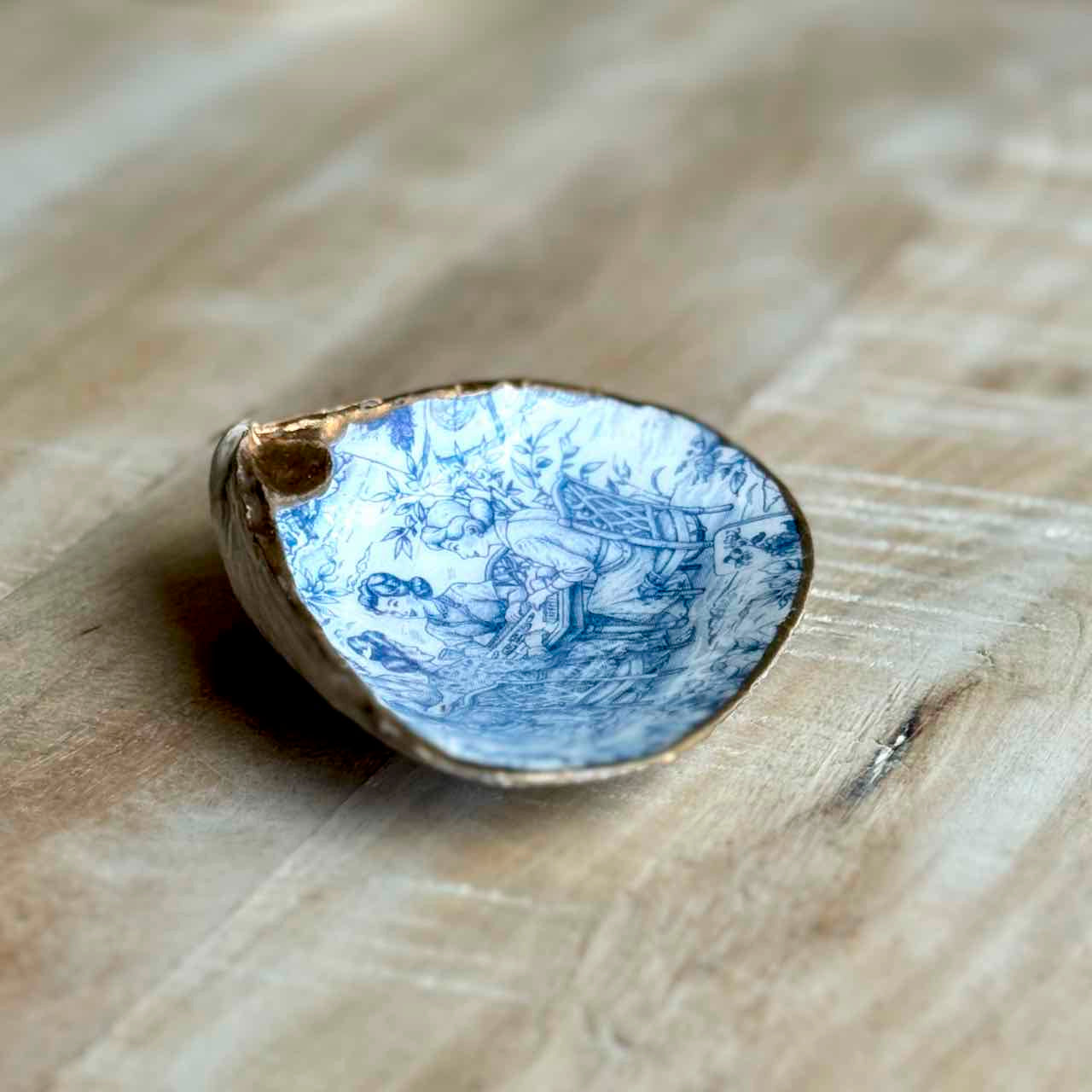 A small ceramic dish with a blue and white illustrated pattern and a gold-toned rim, photographed on a light wood surface.