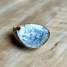 A small ceramic dish with a blue and white illustrated pattern and a gold-toned rim, photographed on a light wood surface.