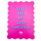A bright pink rectangular mat with wavy scalloped edges, featuring the text 'TILES ARE MY LOVE LANGUAGE' in light blue 3D-style letters. A small white logo for 'MASTONE' is visible at the bottom center.
