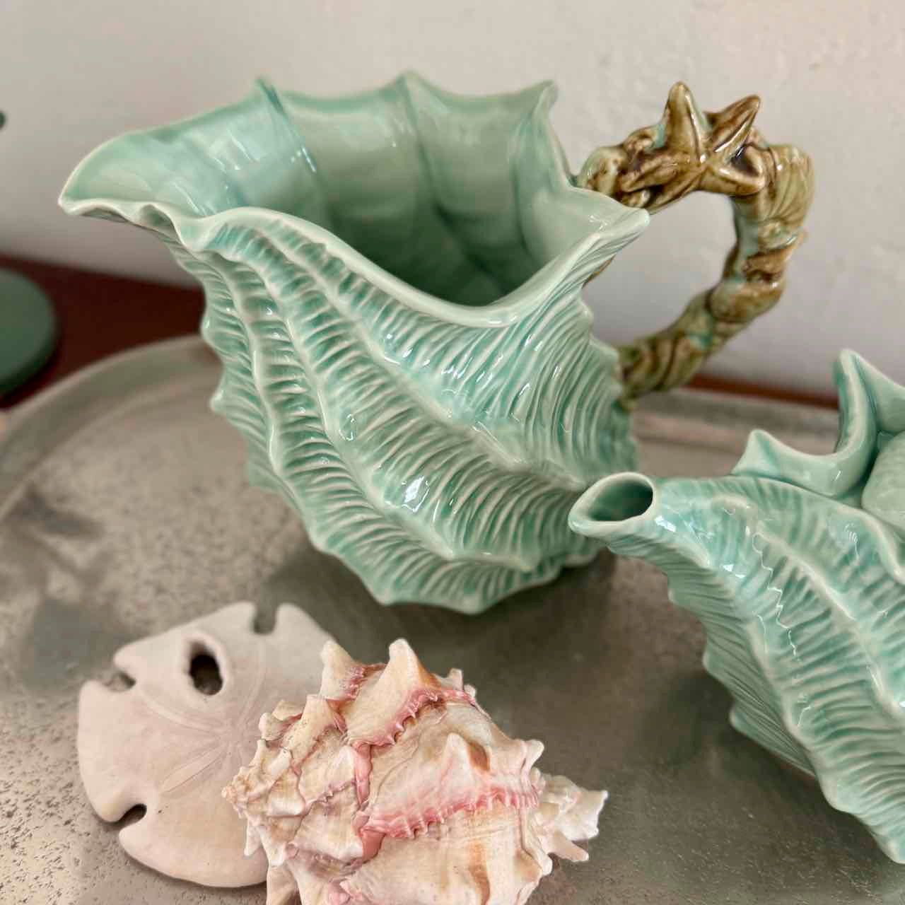 Ceramic vase shaped like a shell with a starfish handle, surrounded by real shells on a neutral background.