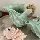 Ceramic vase shaped like a shell with a starfish handle, surrounded by real shells on a neutral background.