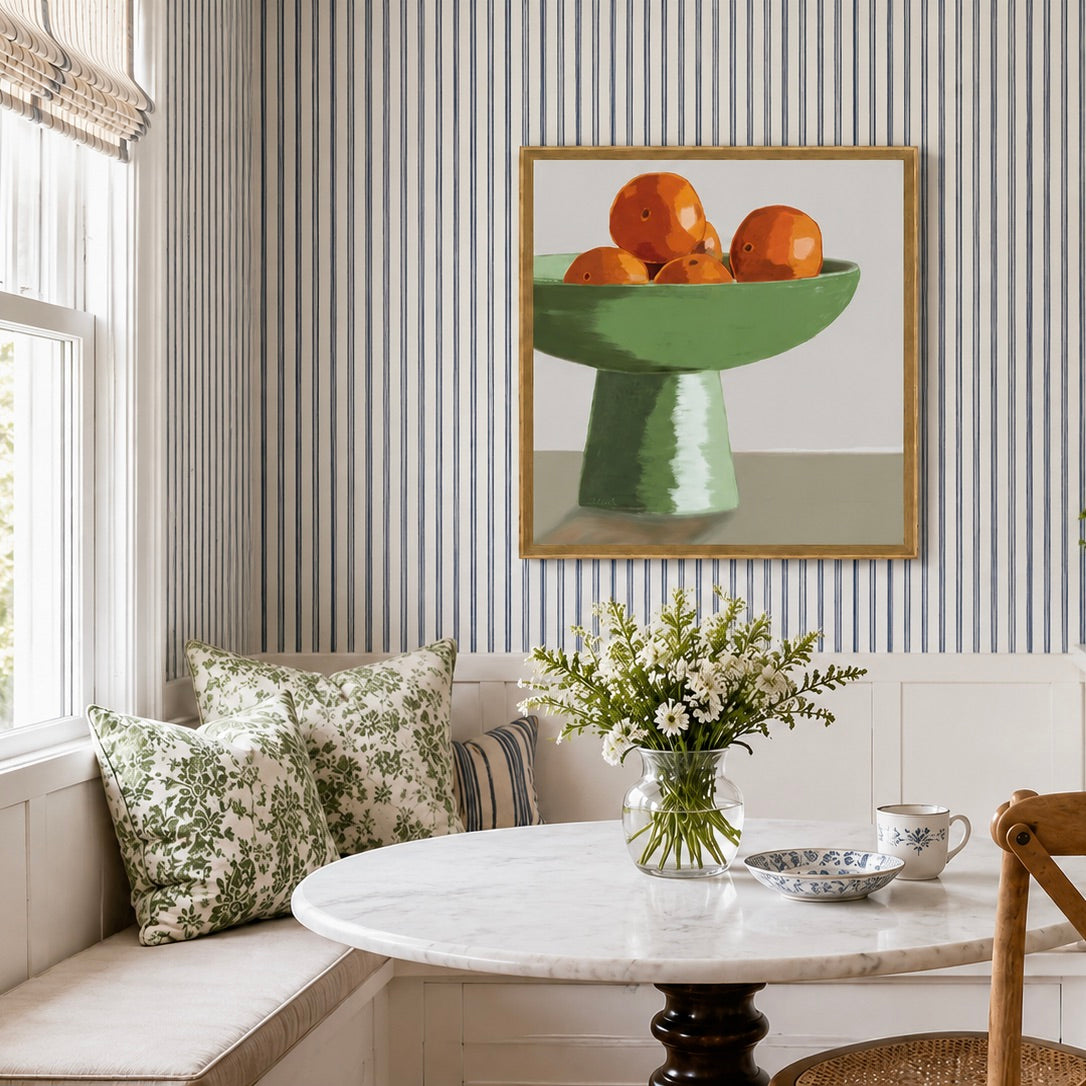 A framed still life painting of oranges in a green pedestal bowl hangs on a striped wall above a window and a round marble table with a floral vase and teacups in a bright dining nook.