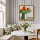 A framed still life painting of oranges in a green pedestal bowl hangs on a striped wall above a window and a round marble table with a floral vase and teacups in a bright dining nook.