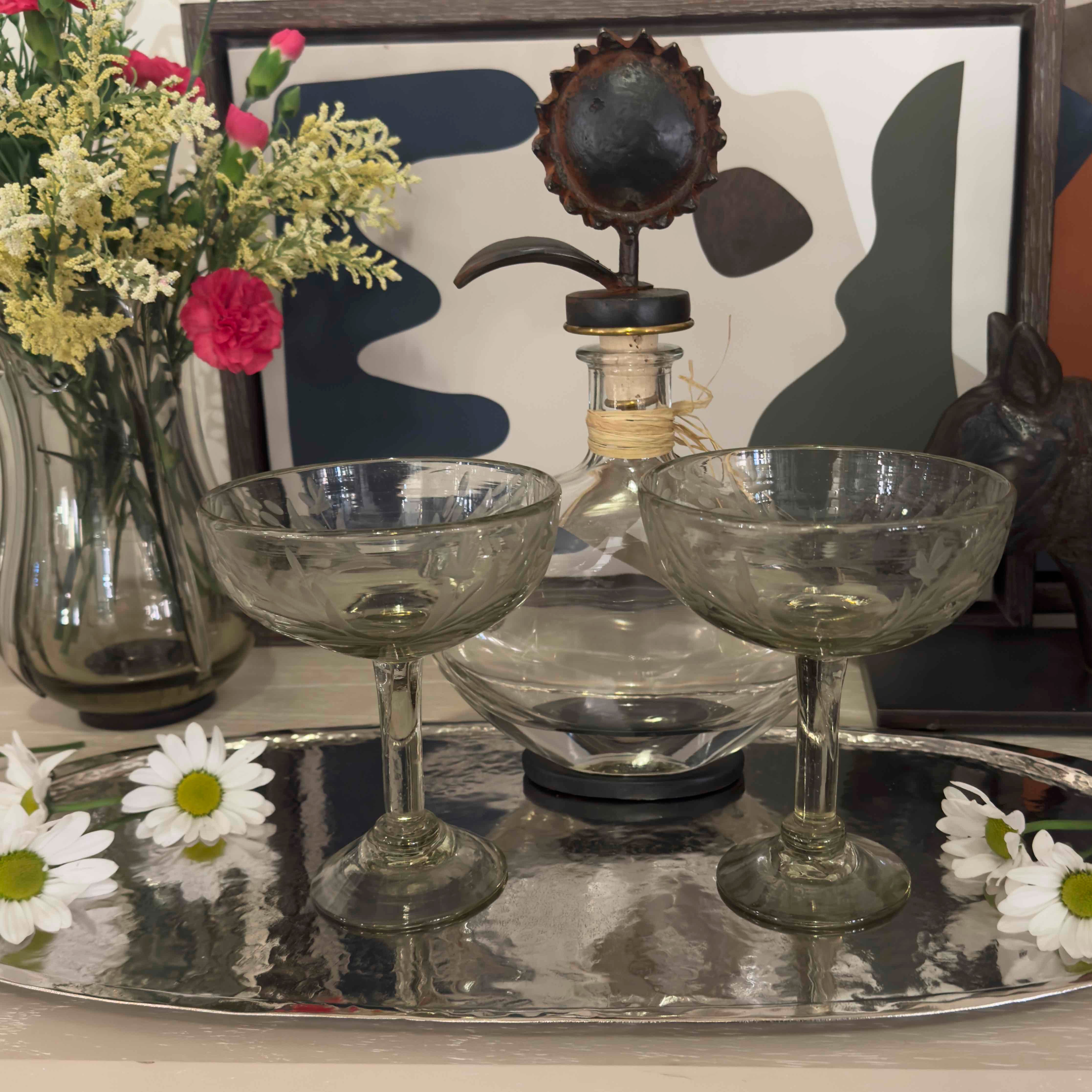 Two clear glass coupes on a silver tray with flowers and decorative items in the background.