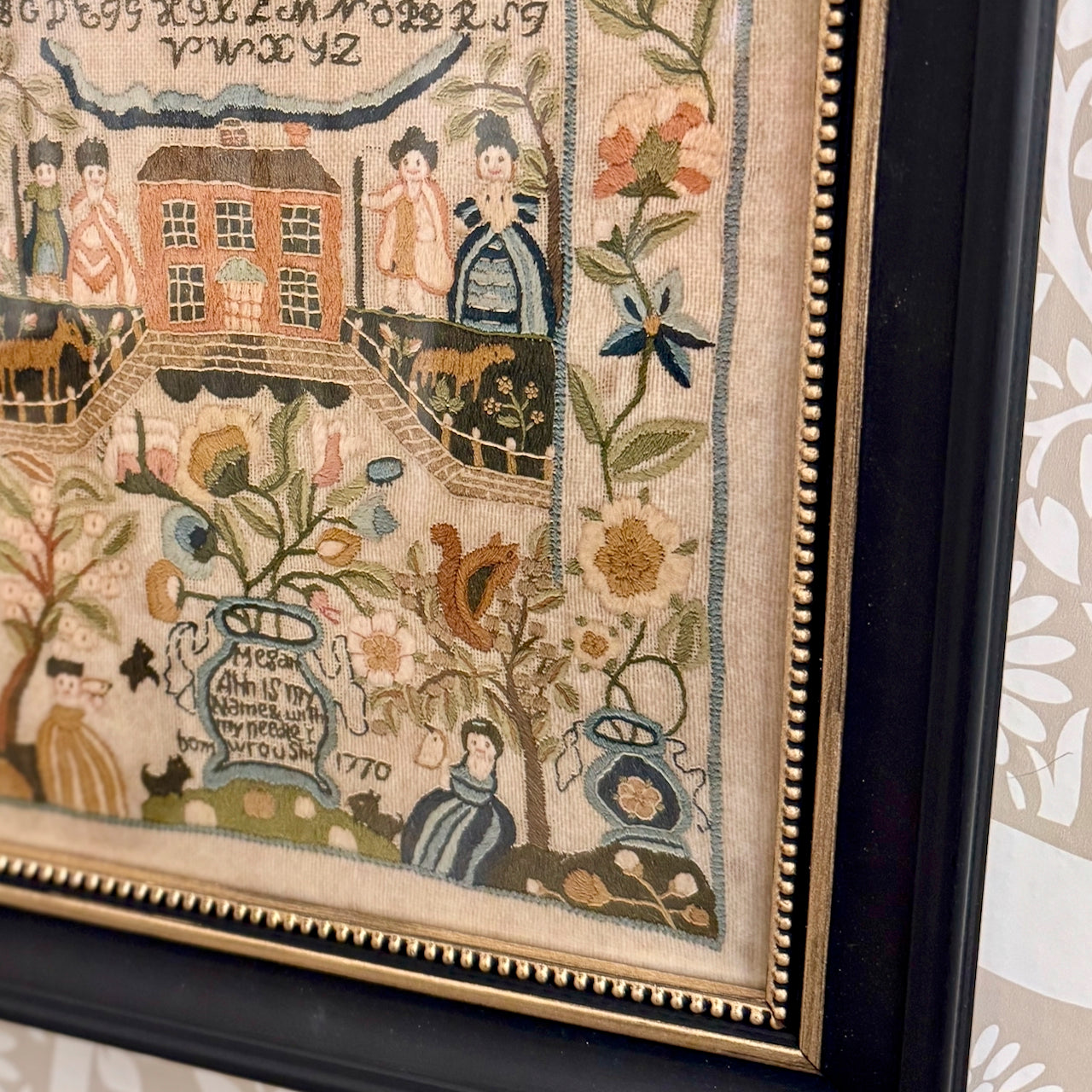 Framed embroidered sampler with intricate designs and text, featuring people, animals, and decorative elements.