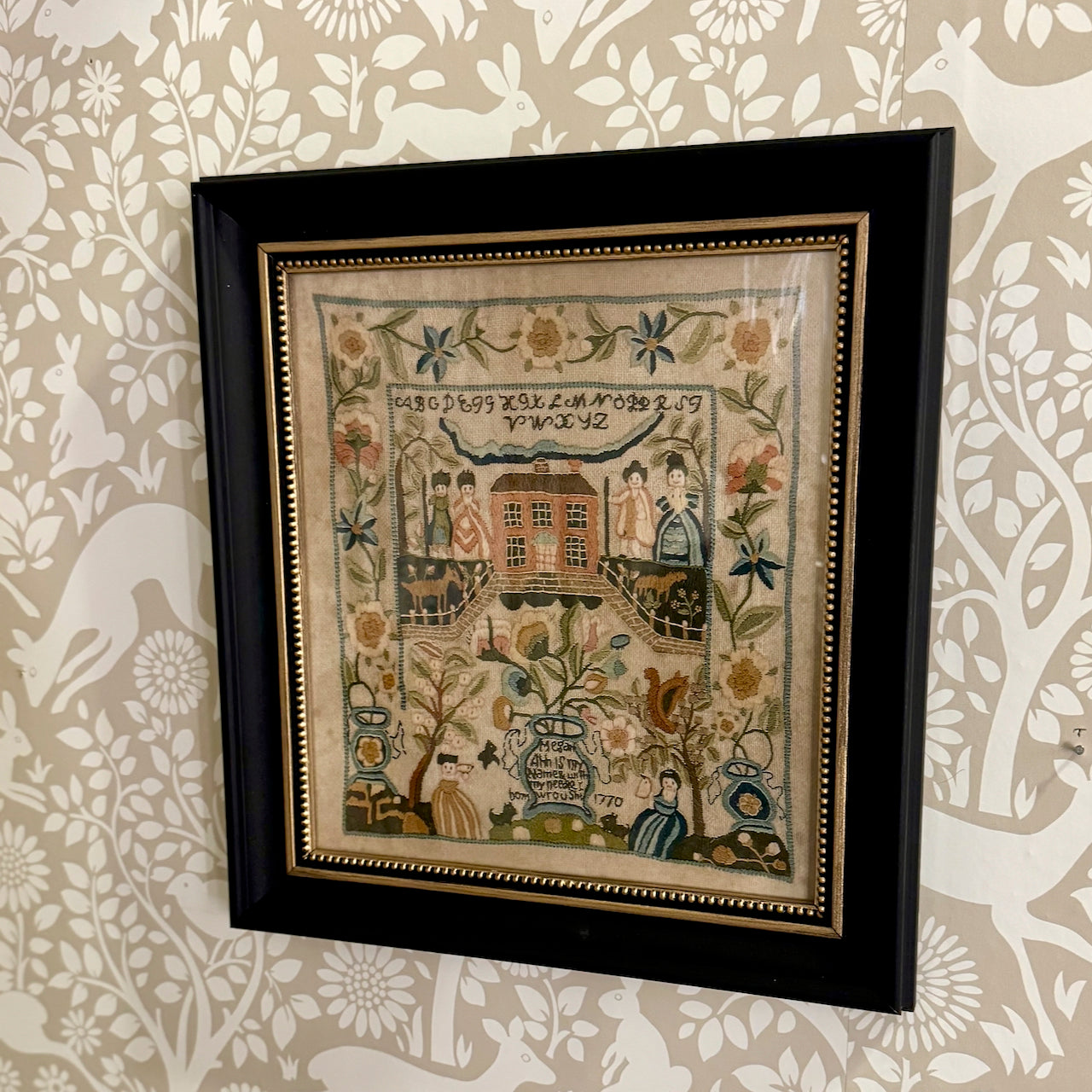 Framed embroidered sampler with decorative elements on a patterned wall.