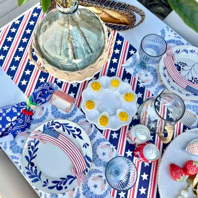 A styled outdoor picnic table setting with patriotic red, white, and blue tableware, including plates with floral and star patterns, napkins, a glass carafe, and a white egg holder with hard-boiled eggs.