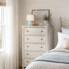 An off-white five-drawer dresser with brass knobs in a bright, neutral bedroom setting next to a bed with white and blue bedding.