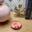 Small pink plate with cherry design on a wooden surface next to a gold decorative item and books.