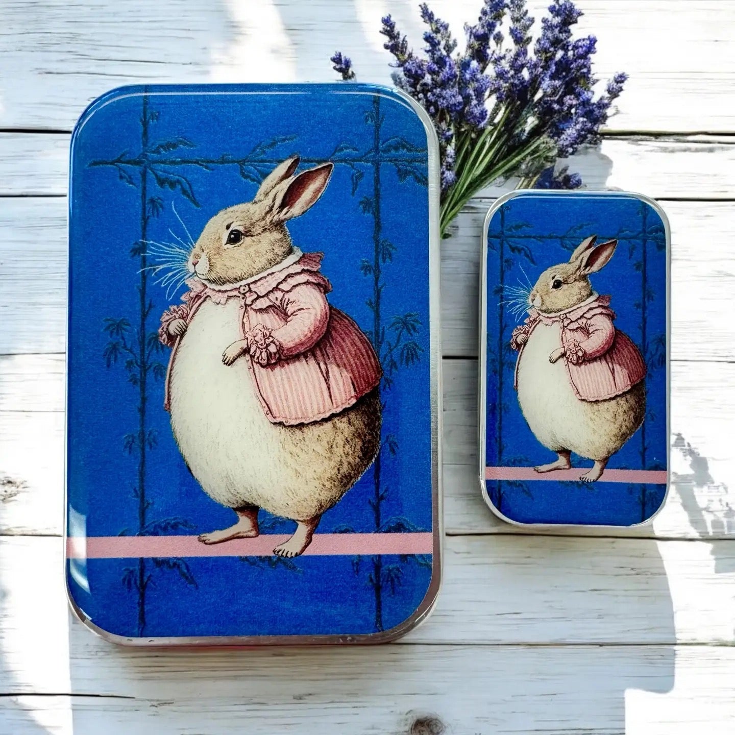 Two tin boxes with a rabbit illustration on a wooden surface with lavender flowers.