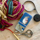 A styled flat-lay of knitting supplies on a light wood surface, featuring a large skein of chunky multicolor yarn, a small tin with a rabbit illustration and metal tools, silver scissors, a wooden crochet hook, and a black circular accessory.