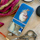 A rectangular metal tin with a blue cover featuring a rabbit illustration, shown open on a wooden surface with sewing tools inside. The tin is surrounded by colorful yarn, silver scissors, and a thimble.