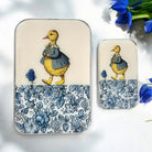 Two rectangular metal tins with rounded corners, one large and one small, featuring a vintage-style illustration of a yellow duckling in a blue dress on a cream background with a blue floral pattern along the bottom. The tins are displayed on a white surface with blue flowers in the corner.