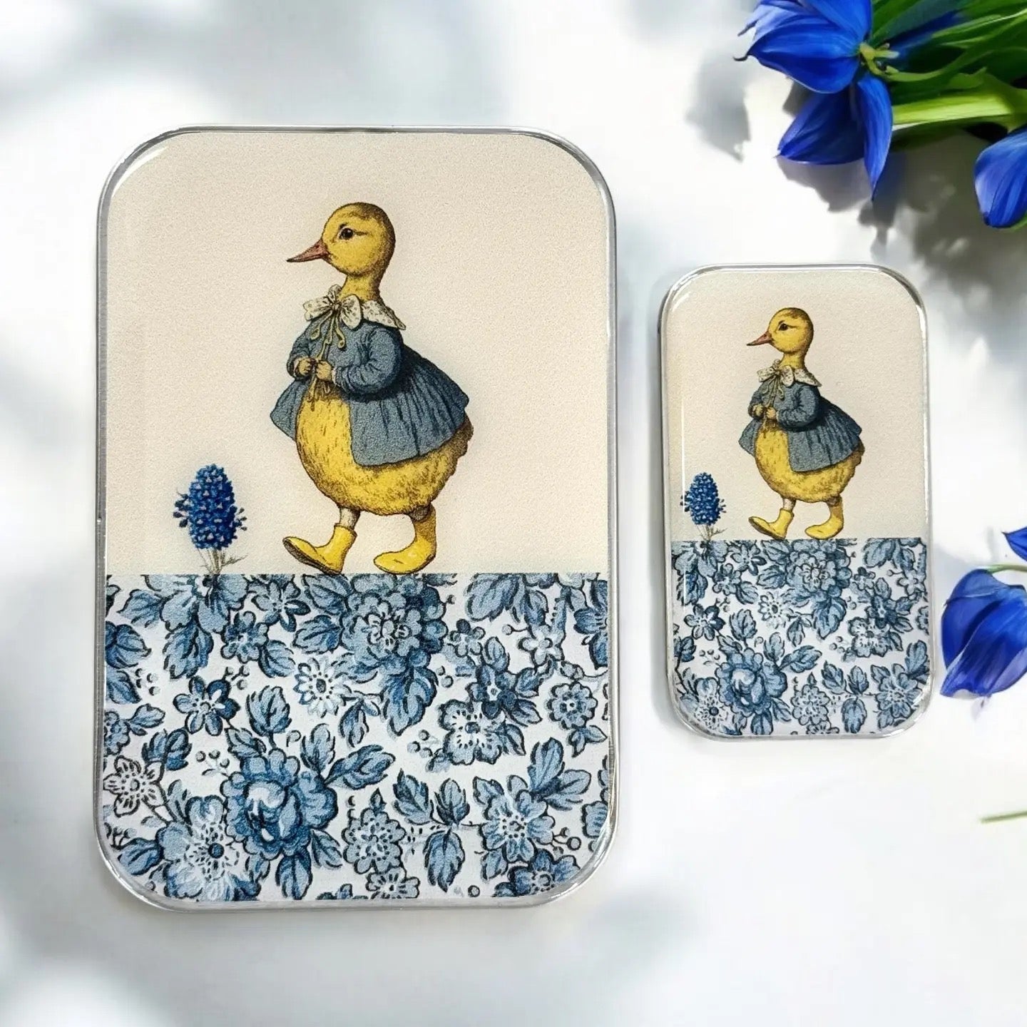 Two rectangular metal tins with rounded corners, one large and one small, featuring a vintage-style illustration of a yellow duckling in a blue dress on a cream background with a blue floral pattern along the bottom. The tins are displayed on a white surface with blue flowers in the corner.