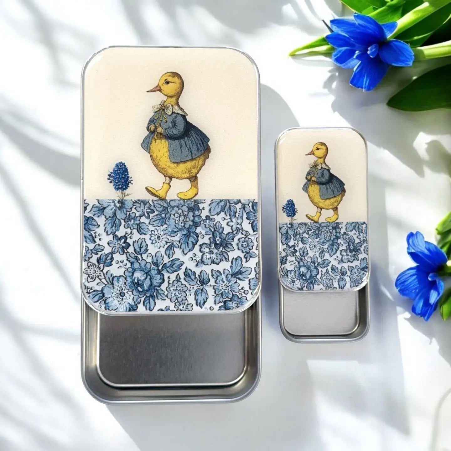 Two tin containers with a duck illustration and floral pattern on a white surface with blue flowers.