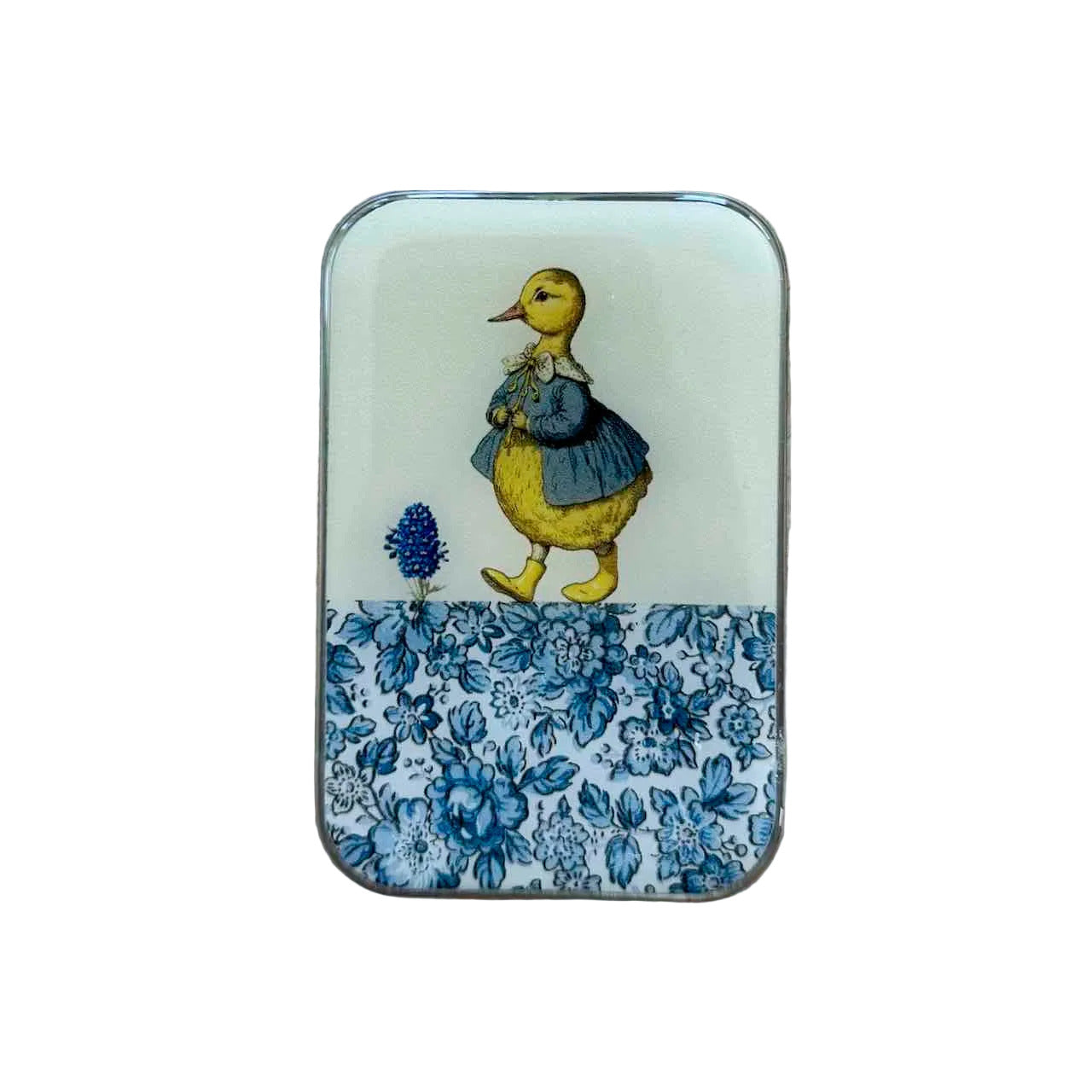 A small rectangular tin with rounded corners, featuring a vintage-style illustration of a yellow duckling in a blue dress on an off-white background, with a blue floral border along the bottom. The tin is photographed from above on a light wood surface.