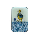 A small rectangular tin with rounded corners, featuring a vintage-style illustration of a yellow duckling in a blue dress on an off-white background, with a blue floral border along the bottom. The tin is photographed from above on a light wood surface.