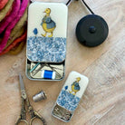 Two decorative metal sewing tools cases with a blue floral pattern and a yellow duck illustration, shown open on a wooden surface with sewing accessories including scissors, thread, pins, and a thimble.