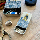 Two decorative metal sewing tools cases with blue floral patterns and a yellow duck illustration, shown open on a wooden surface with sewing accessories like scissors, a thimble, pins, and thread.