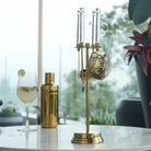 A brass cocktail bar tool set with a stand, strainer, and shaker parts, displayed on a table next to a cocktail shaker and a glass of cocktail in a bright, modern setting.