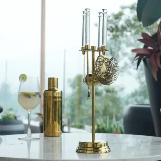 A brass cocktail bar tool set with a stand, strainer, and shaker parts, displayed on a table next to a cocktail shaker and a glass of cocktail in a bright, modern setting.