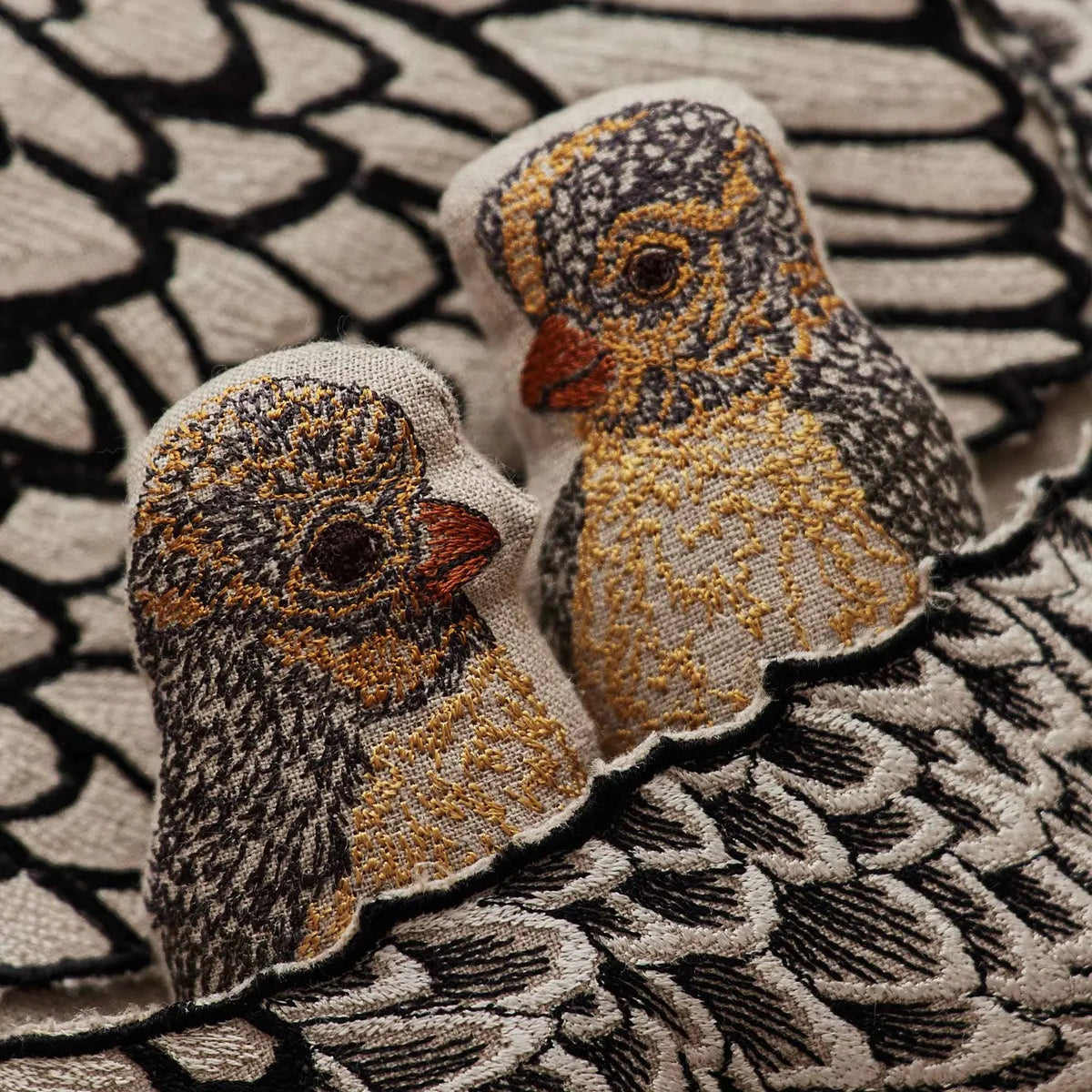 Two small embroidered bird figures with gray, black, and tan stitching rest on a textured woven surface with a black abstract pattern.