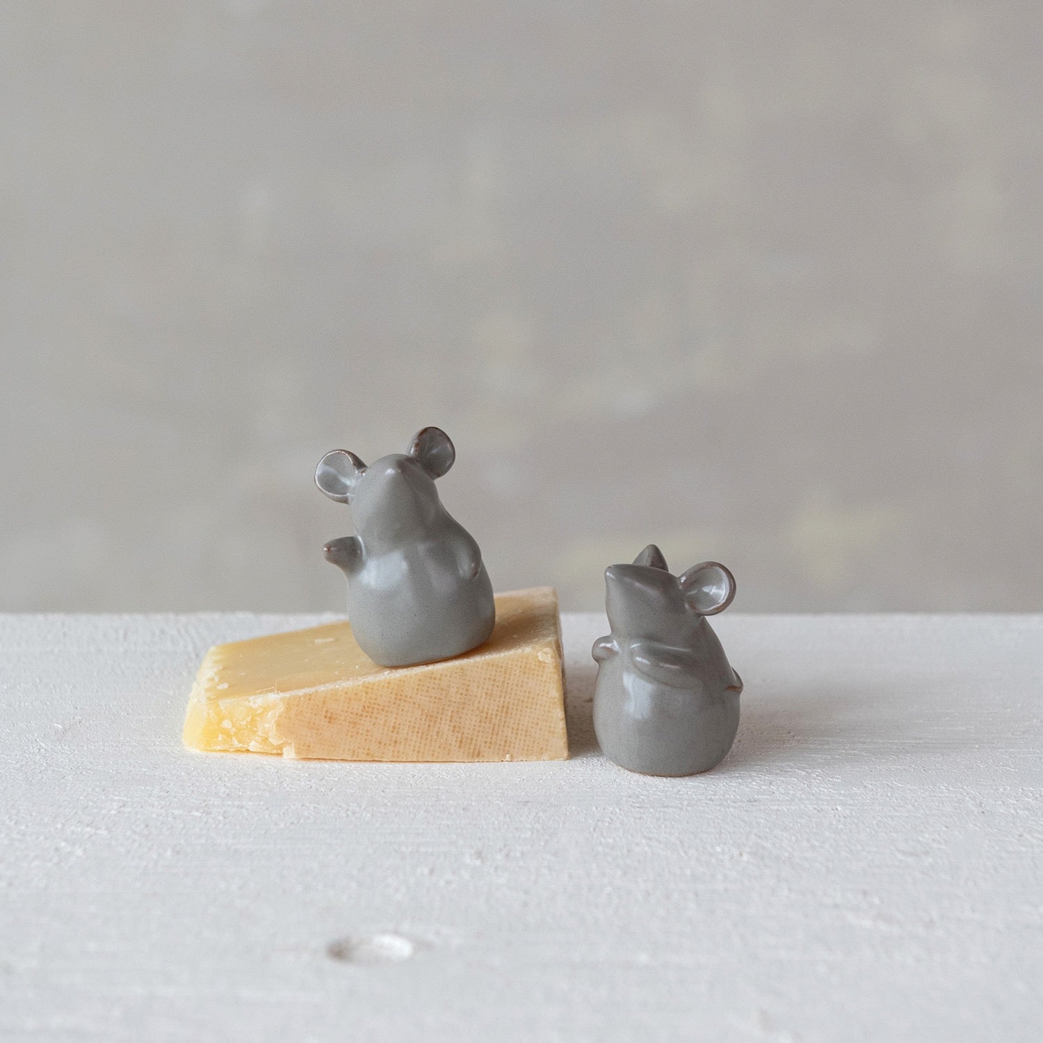 Two small gray ceramic mice on a block of cheese against a plain background