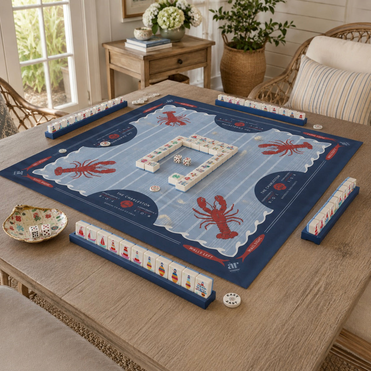 A Mahjong-style game board set arranged on a wooden table in a bright, coastal-themed living room. The board is navy blue with red lobster illustrations and white tile faces, surrounded by matching blue racks and a small decorative bowl.