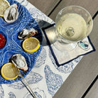 A blue and white oyster platter with lemon slices and a glass of white wine on a patterned placemat on a gray wooden table.