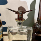 Clear glass decanter with a bronze bull head stopper on a neutral background