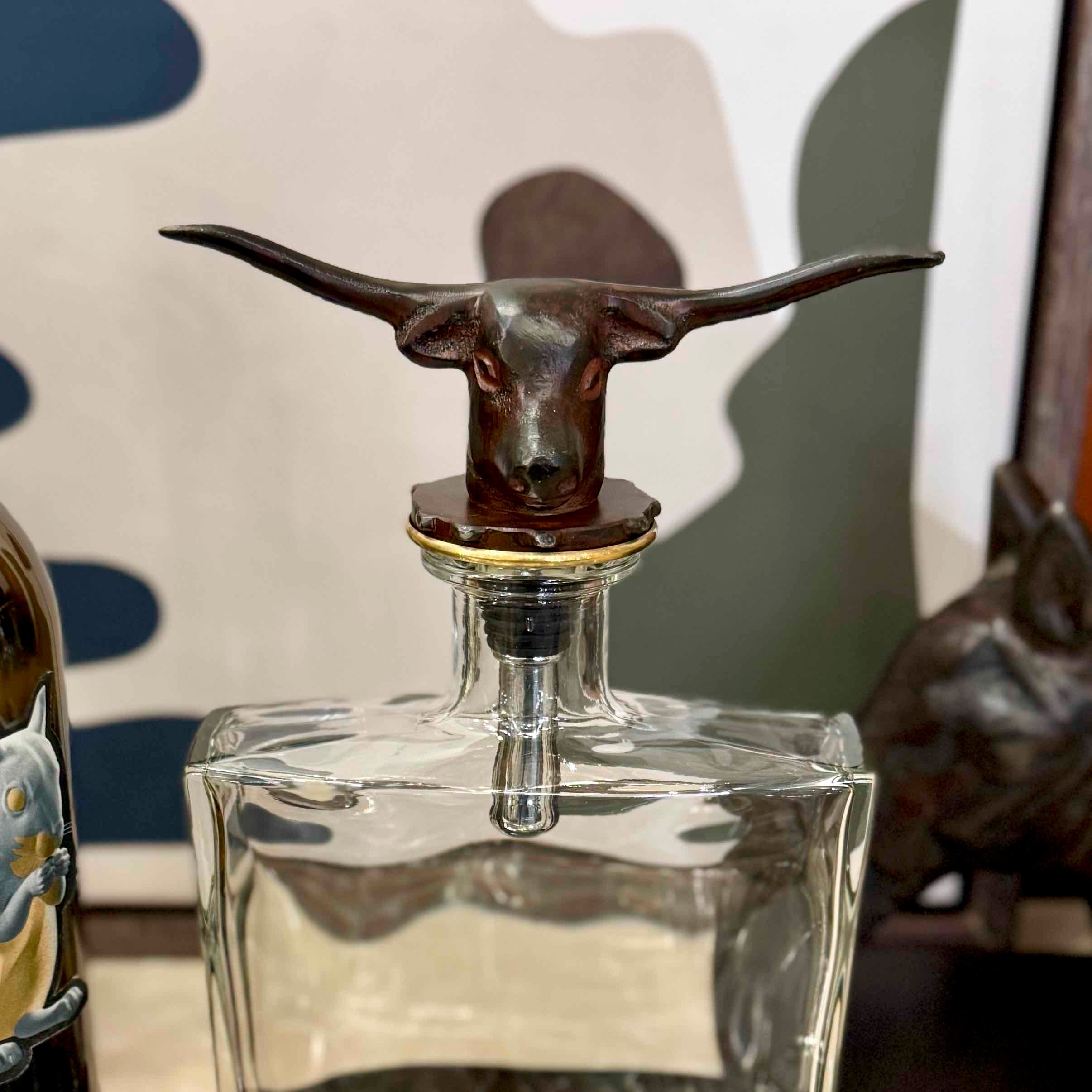 Clear glass decanter with a bronze bull head stopper on a neutral background