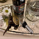 Wine bottle stopper, decorative item, and glass on a metallic tray with flowers.