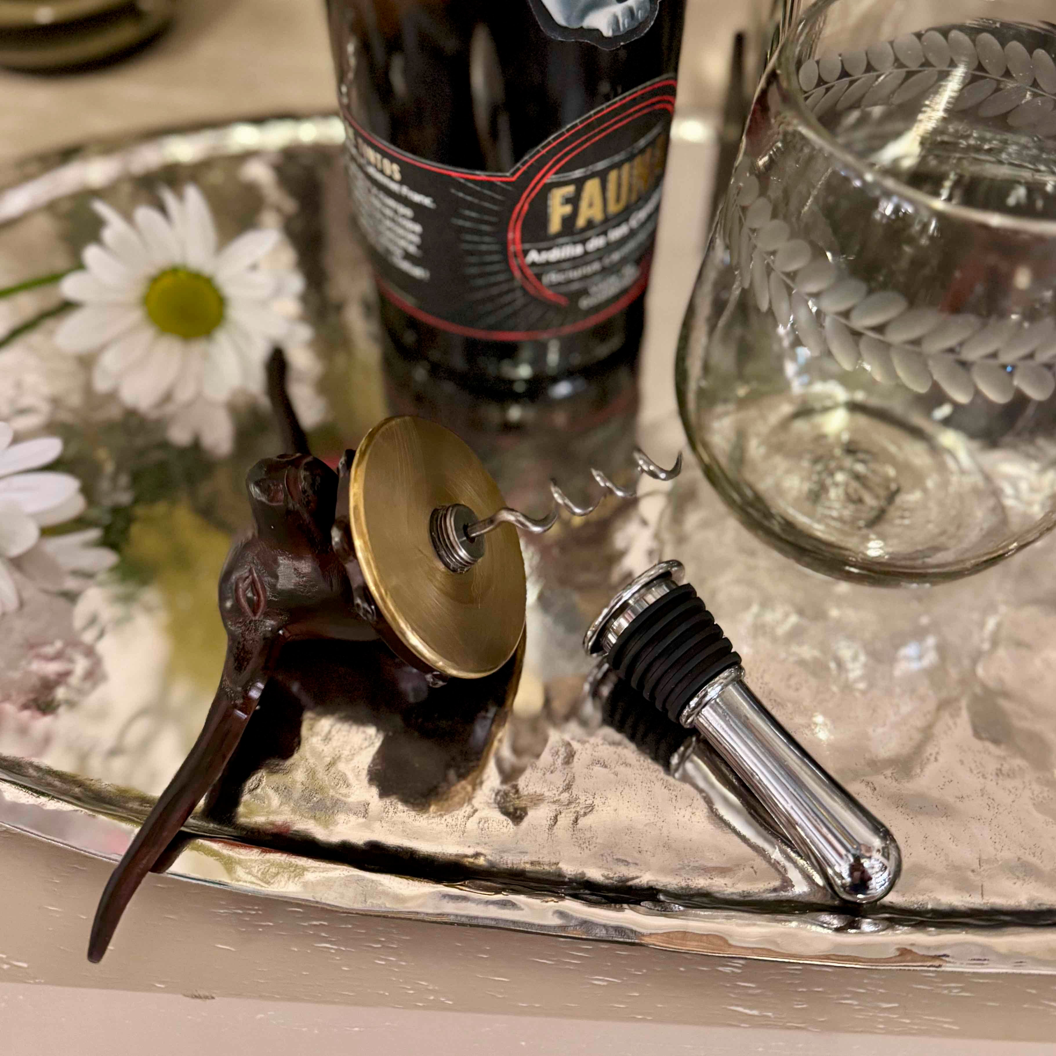 Wine bottle stopper, decorative item, and glass on a metallic tray with flowers.