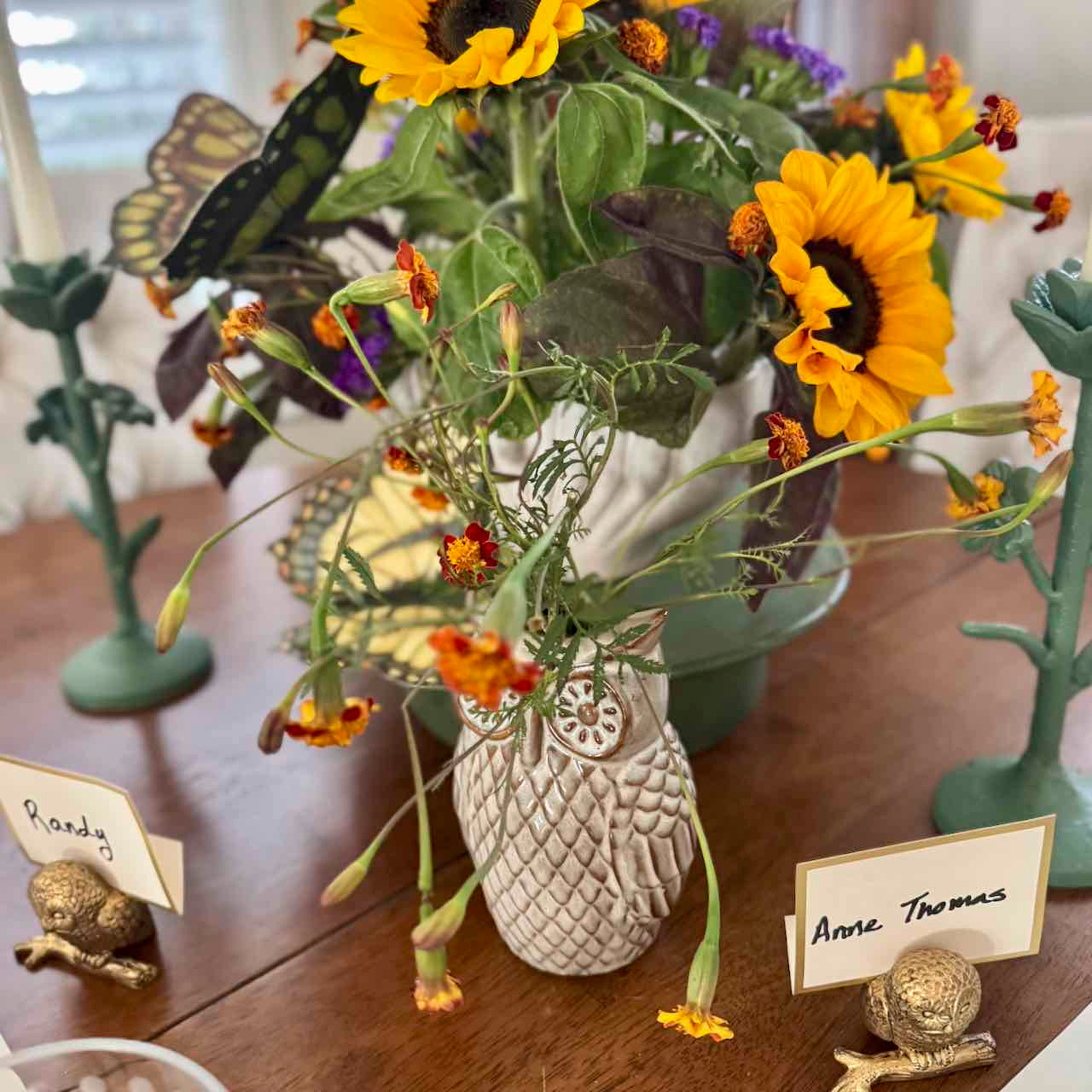 Decorative table setting with a vase of flowers, name cards, and small figurines on a wooden surface.