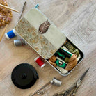 A metal sewing kit tin with a decorative owl illustration on the lid, shown open on a wooden surface with thread spools, scissors, a thimble, and other sewing tools arranged around it.