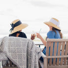Two women sitting on a wooden bench wearing sun hats, enjoying drinks.