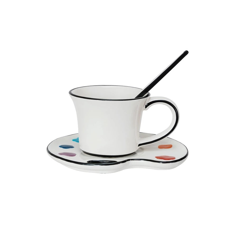 White mug with black rim on a matching saucer with color swatches, accompanied by a black spoon.