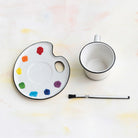 White mug with black rim on a white surface next to a paint palette with colorful dots and a brush.